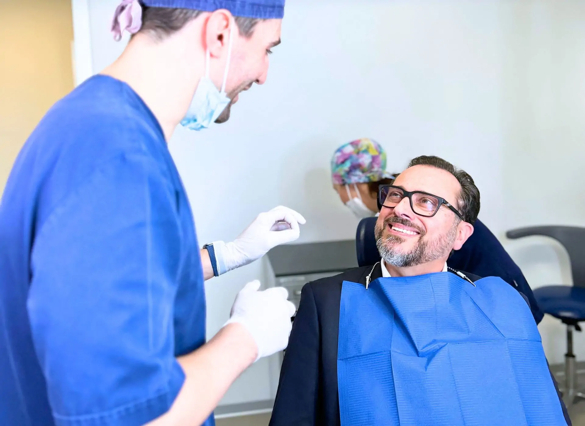 Dentista aperto la domenica a Brescia e provincia 1 Odontoiatra a colloquio con un paziente in poltrona, con l’assistente alle spalle, da Facchinetti Dental a Manerbio